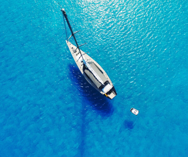 Aerial view of a high-end sailing yacht anchored in turquoise sea with a small tender nearby.