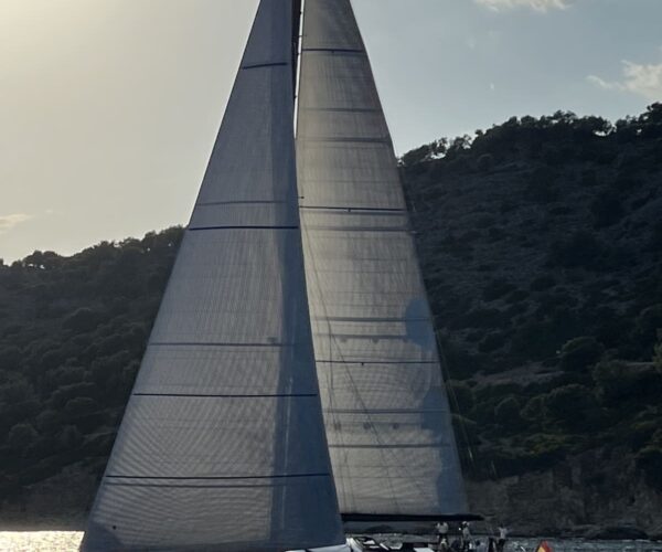 Elegant sailing yacht with tall sails cruising near hillside at sunset.