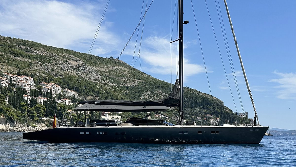 A sleek black sailing yacht anchored in calm waters with scenic mountains in the background.